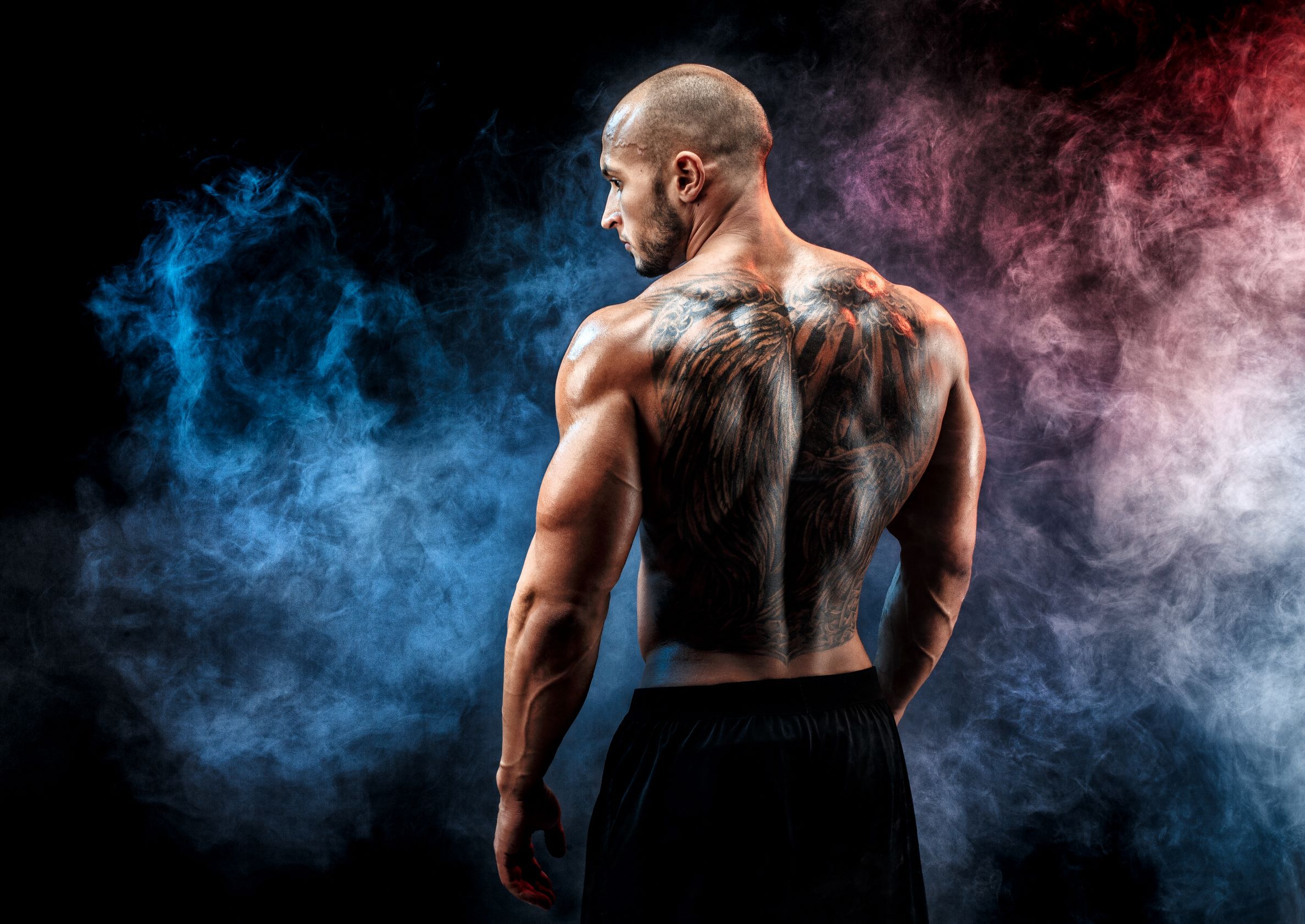 Muscular man with intricate back tattoos, standing against a smoky backdrop, representing themes of body art and aesthetic treatments.